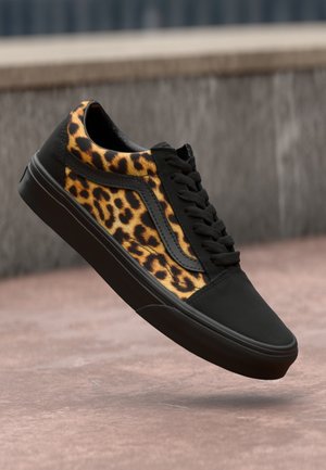Črne teniske z leopardjim vzorcem na panelih, teksturirane tkanine in črnimi vezalkami. Podplat je gumijast in ima raven profil za stabilnost.