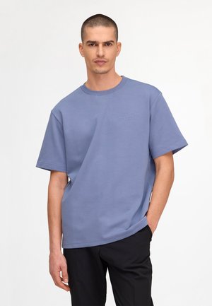 Mężczyzna o krótkich włosach, ubrany w niebieską, luźną koszulę z krótkim rękawem i czarne spodnie, stojący z jedną ręką w kieszeni.