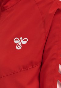 Red fabric jacket section with a small white bee-shaped logo on the chest and white reflective stripes on the sleeve.