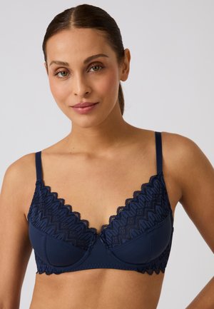 Donna con capelli castani raccolti che indossa un reggiseno in pizzo blu navy con motivi a zigzag e spalline sottili su uno sfondo chiaro uniforme.