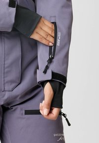 Paarse waterdichte jas met zwarte accenten. Bevat een veilige zipzak op de mouw en een verstelbare klittenbandsluiting bij de manchetten. Tekst: "STY™ 8000."