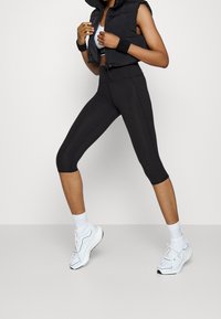 Czarne legginsy o kroju przed kostkę w połączeniu z czarną, bezrękawnikową kurtką puchową. Białe buty sportowe i białe skarpetki sięgające kostek dopełniają outfit.