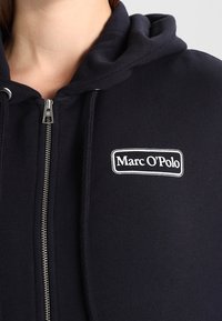 Felpa con zip di colore navy realizzata in morbido cotone. Presenta un'applicazione con logo bianco e cappuccio con cordino, con una texture liscia e zip metallica.