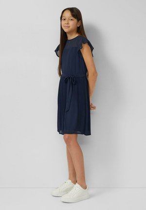 Marineblaues, plissiertes Kleid mit kurzen, gerafften Ärmeln und gebundener Taille, kombiniert mit weißen Turnschuhen. Glatter Stoff und knielanger Schnitt.