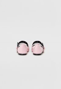 Růžové lakované baletní boty s černým páskem a přezkou, vyznačující se zaoblenou špičkou a plochou podrážkou.