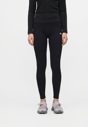 Personne portant un legging noir taille haute avec le logo Adidas et des chaussures de sport grises, debout devant un fond blanc uni.
