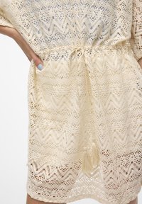 Abito in pizzo color crema con motivo a zigzag, presenta una vita elasticizzata e dettagli con frange, realizzato in un tessuto leggero e traspirante.