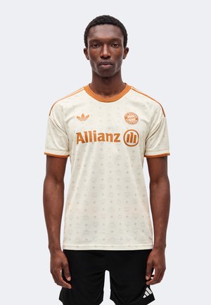 Un mannequin masculin se tient debout face à l’avant, vêtu d’un maillot de football crème du Bayern Munich avec des finitions orange et des logos de sponsors, assorti à un short noir.