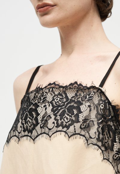 Κοντινό πλάνο γυναίκας που φοράει μπεζ τοπ με μαύρη δαντέλα με λουλούδια και λεπτές μαύρες τιράντες πάνω σε απλό φόντο.