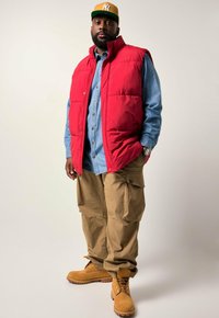 Rote gepolsterte Weste über einem Denimhemd, khakifarbene Cargo-Hosen und braune Stiefel. Zu den Accessoires gehören eine gelbe Mütze und eine Uhr. Schlicher Hintergrund.