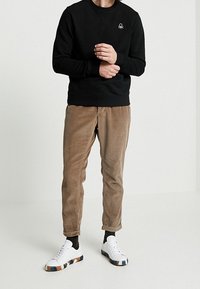 Svart sweatshirt med en liten logotyp, tillsammans med ljusbruna kordbyxor och vita sneakers med en flerfärgad sula.