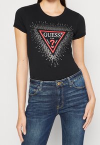 Camiseta de algodón negra con un logotipo en forma de triángulo rojo con acentos de puntos plateados y texto, combinada con unos jeans de mezclilla azul oscuro.