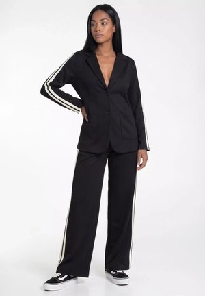 Kobieta ubrana w czarny garnitur z białymi paskami na rękawach i spodniach, w połączeniu z czarno-białymi trampkami, stoi na białym tle.