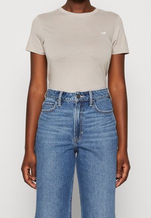 Vaaleanbeige lyhythihainen t-paita pyöreällä pääntiellä ja pienellä logolla, yhdistettynä korkeavyötäröisiin sinisiin farkkuihin, joissa on klassinen leikkaus ja ompelukoristeita.