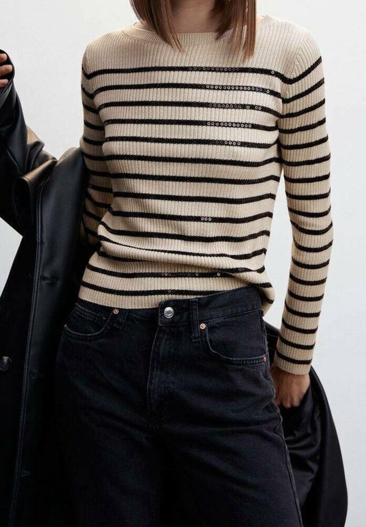 Maglione a coste beige con strisce orizzontali nere e dettagli in paillettes, abbinato a jeans in denim neri a vita alta.