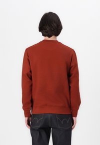 Roestkleurige sweatshirt met een ronde hals, lange mouwen en een solide textuur. Voorzien van geribde manchetten en onderrand, gecombineerd met donkere spijkerbroeken.