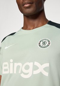 Maillot de sport vert clair avec un col rond, des accents noirs sur les épaules, un grand logo "Bingx" blanc et un badge du Chelsea FC. Tissu texturé.