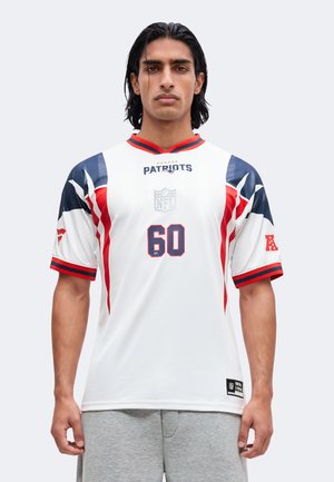 Mężczyzna z ciemnymi włosami do ramion nosi biały jersey New England Patriots NFL z numerem 60 i szare spodenki na jednolitym tle.