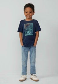 Navyblaues T-Shirt mit einem geometrischen Blätter-Design in Hellblau und Koralle. Kombiniert mit hellblauen Jeans und weißen Sneakern.