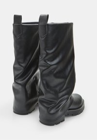 Un paio di stivali alti in pelle nera con linguette per la presa, punte arrotondate e suole in gomma spessa, posizionati in verticale su una superficie bianca.