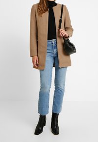 Žena v camel kabátu, černém roláku, světle modrých zkrácených džínách, černých podpatkových botách a s černou ramenní taškou.