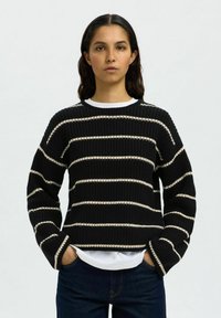 Donna con i capelli scuri che indossa un maglione nero a coste con strisce orizzontali bianche e jeans scuri, mani in tasca, espressione neutra.