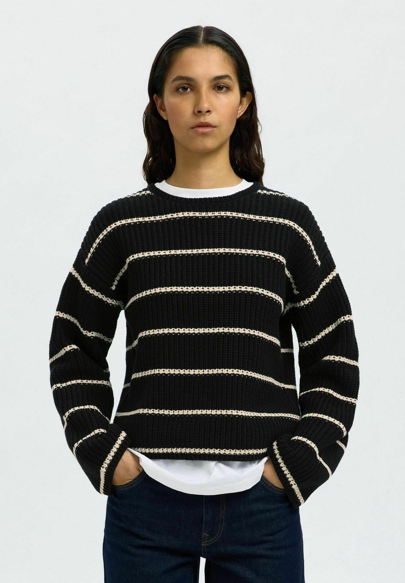 Donna con i capelli scuri che indossa un maglione nero a coste con strisce orizzontali bianche e jeans scuri, mani in tasca, espressione neutra.