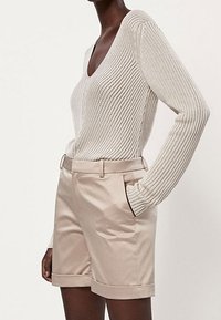 Beige gerippter Strickpullover mit V-Ausschnitt, kombiniert mit hellbeigen maßgeschneiderten Shorts mit seitlichen Taschen und umgeschlagenen Säumen.