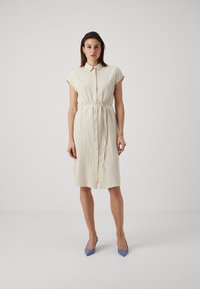 Rochie cămașă bej deschis cu mâneci scurte, închidere cu nasturi în față, talie cu curea și țesătură texturată. Asociată cu pantofi cu toc ascuțit, de culoare albastră.