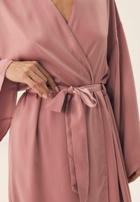 Rękawiczka w stylu kimono wykonana z miękkiego różowego satynowego materiału, z szerokimi rękawami i przewiązanym w pasie paskiem. Gładka faktura z minimalnymi detalami szycia.