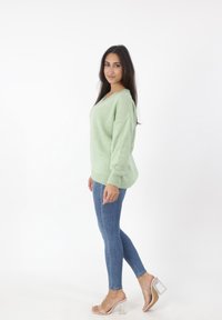 Lichtgroene, gestructureerde trui met V-hals, gedragen met strakke blauwe jeans en transparante blokhak-sandalen, staande op een witte achtergrond.