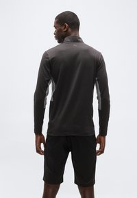 Άνδρας με μαύρο αθλητικό μακρυμάνικο πάνω μέρος με λογότυπο Puma στην άνω πλάτη και μαύρο σορτς, γυρισμένος με την πλάτη σε απλό φόντο.