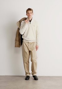Pulover bej deschis cu guler cu fermoar pe jumătate, asortat cu pantaloni lărguți bej și pantofi negri slip-on. O jachetă maro este aruncată pe braț.