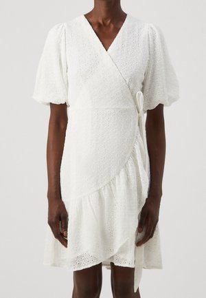 Mørkhudet person iført en hvid øjletslagsvikle kjole med pufærmer og V-udskæring mod en ensfarvet baggrund.