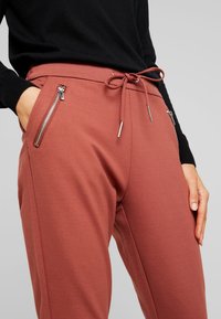 Joggingbroek in roestkleur met een trekkoord in de taille, ritssluitingen aan de zijkanten en een gladde textuur. Gestyled met een zwart topje met lange mouwen.