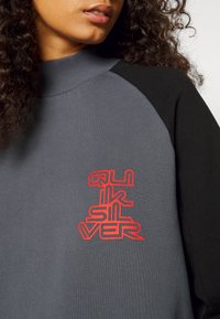 Grå fleece-tröja med svarta ärmar, med en framträdande röd logotyp på bröstet och hög krage. Mjuk textur och avslappnad design.