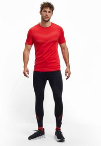 Rotes sportliches T-Shirt mit strukturierten Streifen, kombiniert mit schwarzen Leggings mit roten Akzentlinien. Modell trägt schwarze Turnschuhe.