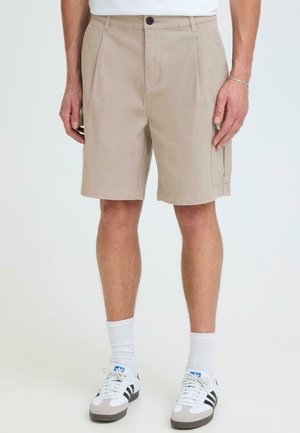 Beige knielange Shorts mit vorderen Falten, getragen von einer stehenden Person, kombiniert mit weißen Socken und weißen Adidas-Sneakern mit schwarzen Streifen.
