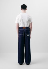 Asmuo su trumpais garbanotais plaukais, vilkintis baltą marškinėlį ir tamsius plačius džinsus su dviem galiniais kišenėmis, iš kurių viena pažymėta "Dickies".