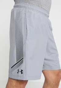 Ljust grå sportshorts med elastisk midja, sidofickor och en svart geometrisk design på vänster ben som har en logotyp.