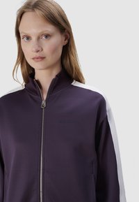 Paarse zip-up jack met een staande kraag, met witte accenten op de schouders en een reliëflogo op de borst. Soepele stoftextuur.
