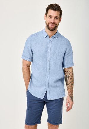 Smiling man with beard wearing a light blue short-sleeve button-up shirt and navy blue shorts, showing tattoos on his right forearm.
