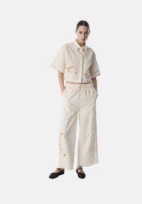 Beige Hemd mit kurzen Ärmeln und aufwendigen Ausschnitten, kombiniert mit weiten beigen Hosen mit ähnlichen Akzenten und schwarzen Flachschuhen.