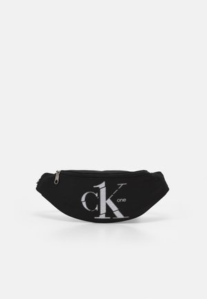 Sort stof bæltetaske med lynlås og hvidt "ck one" logo foran i midten mod ensfarvet baggrund.