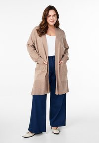 Cárdigan largo beige con frente abierto, dos bolsillos, llevado sobre una blusa blanca y pantalones vaqueros de pierna ancha azul marino. Textura suave, diseño minimalista.