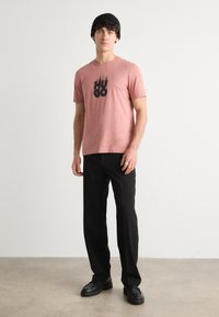 Rīsa piedurkņu rozā T-krekls ar melno "HUGO" logo, saskaņots ar melnām biksēm un melnā apavu pāri. Modelis valkā melnu cepuri. Vienkāršs dizains.