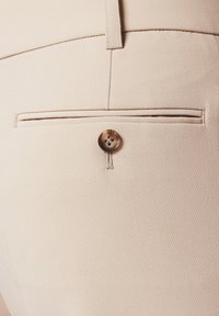 Beżowe materiałowe spodnie mają tylną kieszeń z brązowym guzikiem, teksturowaną powierzchnię oraz dopasowany krój z delikatnym diagonalnym wzorem.