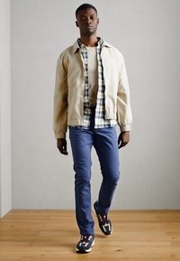 Jasnobeżowa kurtka bombka na checked koszuli, zestawiona z granatowymi spodniami i wielokolorowymi trampkami. Minimalistyczne tło.