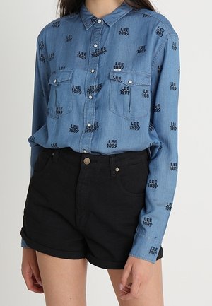 Mujer con camisa azul de botones con texto negro "LEE 1889" y shorts negros de talle alto con dobladillo vuelto.