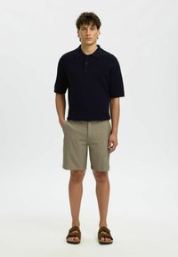 Jeune homme debout, les mains dans les poches, portant un polo noir à manches courtes, un short beige et des sandales marron sur un fond blanc.